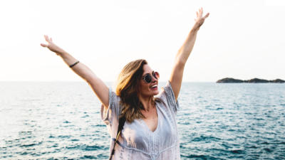 Woman with arms raised over head, sunglasses on, smiling, with ocean in the backgronud